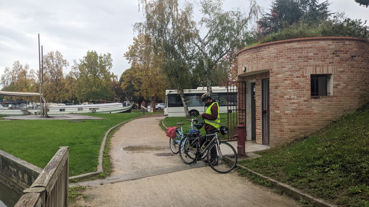 Two bikes and one cyclist outside round washrooms in Castelsarrasin.